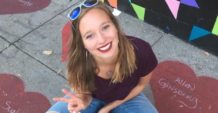 Scopri AuPairCare La ragazza alla pari è seduta per terra, fa il segno della pace e sorride alla telecamera.